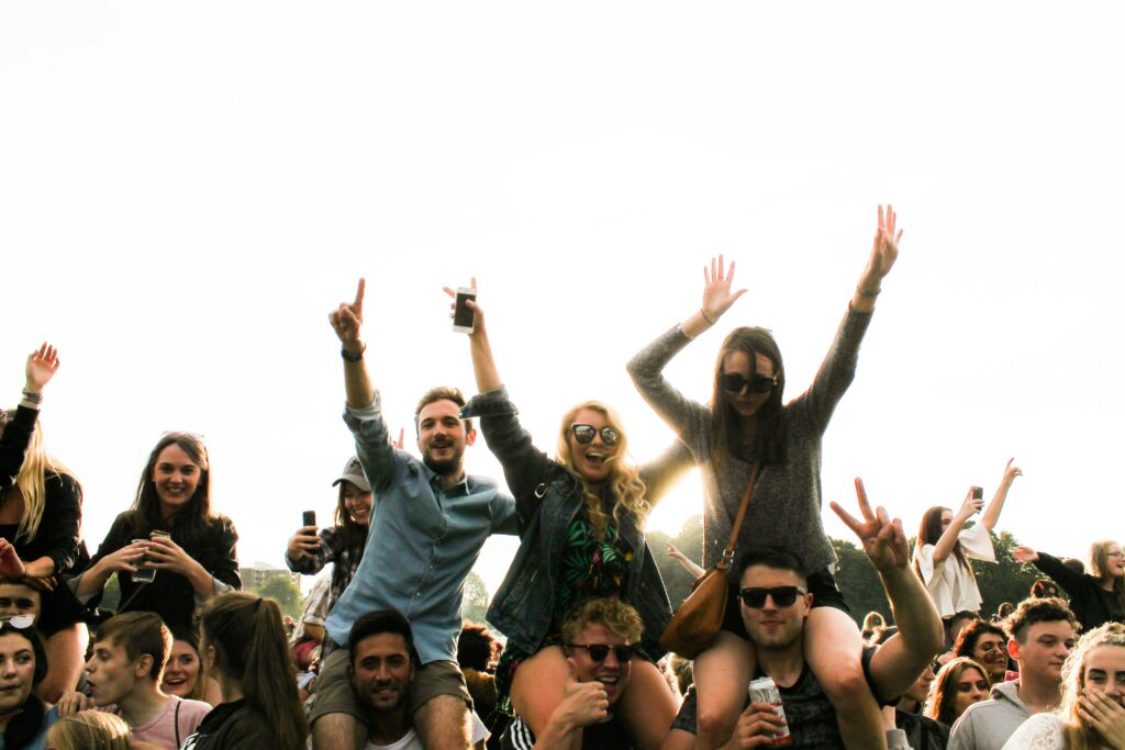 Groupe de jeunes amis souriants et joyeux sur les épaules, levant les bras en l'air lors d'un concert en plein air ou d'un festival de musique événementiel. Scène de foule animée avec des festivaliers célèbrant l'ambiance et la musique, idéal pour illustrer la vie nocturne, un concert ou un événement culturel en extérieur. Les participants sont radieux et l'ambiance festive.